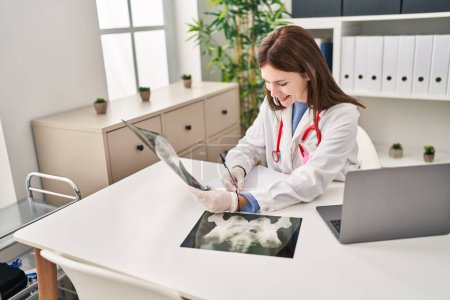 Foto de Mujer joven rubia médico buscando rayos X escribir informe médico en la clínica - Imagen libre de derechos