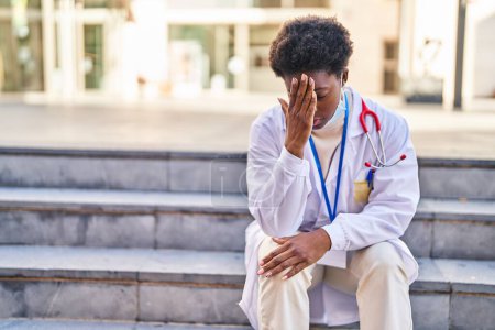 Téléchargez les photos : Femme afro-américaine portant un uniforme de médecin et un masque médical avec une expression inquiète dans la rue - en image libre de droit
