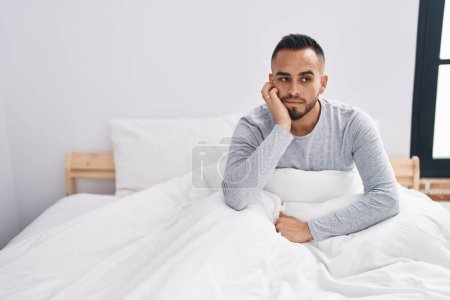 Téléchargez les photos : Jeune homme hispanique couché sur le lit avec une expression sérieuse dans la chambre - en image libre de droit