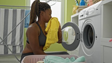 Téléchargez les photos : Femme afro-américaine laver les vêtements sentant t-shirt sale à la buanderie - en image libre de droit
