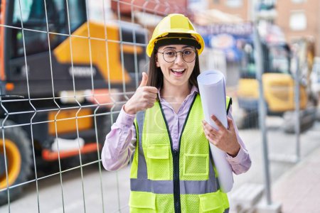 Photo for Hispanic girl wearing architect hardhat at construction site smiling happy and positive, thumb up doing excellent and approval sign - Royalty Free Image