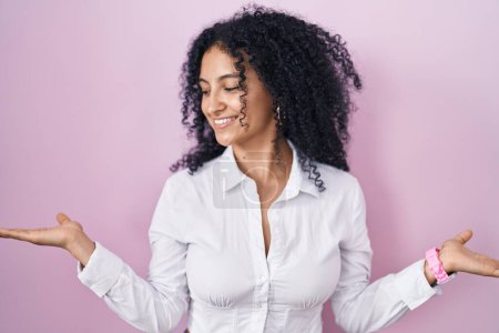 Foto de Hispanic woman with curly hair standing over pink background smiling showing both hands open palms, presenting and advertising comparison and balance - Imagen libre de derechos