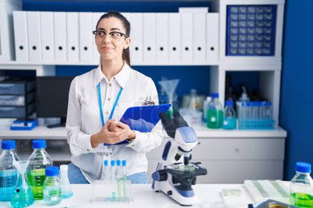 Téléchargez les photos : Jeune femme brune travaillant au laboratoire scientifique souriant regardant vers le côté et regardant loin en pensant. - en image libre de droit