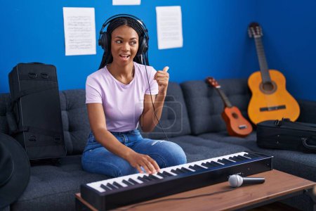 Photo for African american woman with braids playing piano keyboard at music studio pointing thumb up to the side smiling happy with open mouth - Royalty Free Image