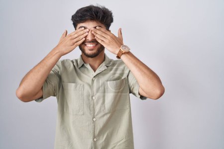 Téléchargez les photos : Homme arabe avec barbe debout sur fond blanc couvrant les yeux avec les mains souriantes gaies et drôles. concept aveugle. - en image libre de droit