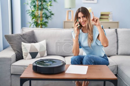 Foto de Mujer hispana joven sentada en casa por robot de vacío hablando por teléfono sorprendida con una idea o pregunta apuntando con el dedo con la cara feliz, número uno - Imagen libre de derechos