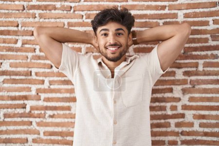 Téléchargez les photos : Homme arabe avec barbe debout sur fond de mur de briques relaxant et étirant, bras et mains derrière la tête et le cou souriant heureux - en image libre de droit