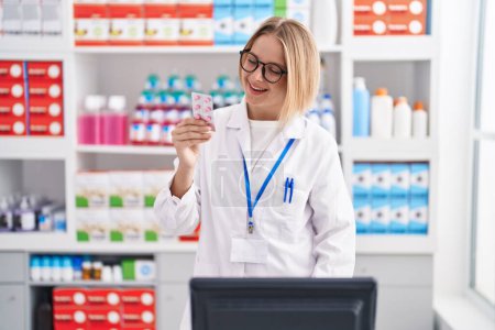 Téléchargez les photos : Jeune femme blonde pharmacien souriant confiant tenant comprimés comprimés à la pharmacie - en image libre de droit