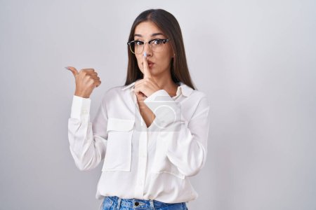 Photo for Young brunette woman wearing glasses asking to be quiet with finger on lips pointing with hand to the side. silence and secret concept. - Royalty Free Image
