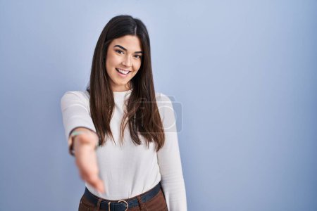 Foto de Mujer morena joven de pie sobre fondo azul sonriendo amistoso ofreciendo apretón de manos como saludo y bienvenida. negocios exitosos. - Imagen libre de derechos