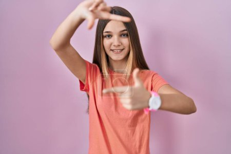 Foto de Chica adolescente de pie sobre fondo rosa sonriendo haciendo marco con las manos y los dedos con la cara feliz. concepto de creatividad y fotografía. - Imagen libre de derechos