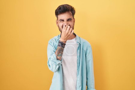 Téléchargez les photos : Young hispanic man with tattoos standing over yellow background smelling something stinky and disgusting, intolerable smell, holding breath with fingers on nose. bad smell - en image libre de droit