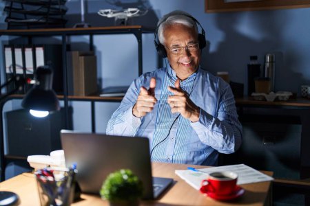Photo for Hispanic senior man wearing call center agent headset at night pointing fingers to camera with happy and funny face. good energy and vibes. - Royalty Free Image