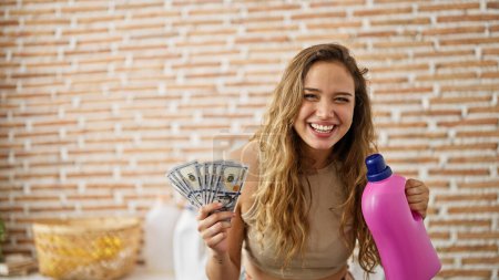 Photo for Young beautiful hispanic woman holding detergent bottle and dollars at laundry room - Royalty Free Image