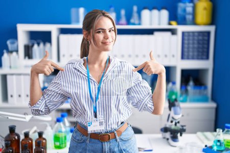 Foto de Young beautiful woman working at scientist laboratory looking confident with smile on face, pointing oneself with fingers proud and happy. - Imagen libre de derechos