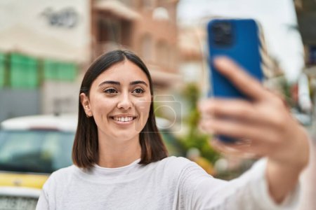 Foto de Mujer hispana joven sonriendo confiada haciendo selfie por el teléfono inteligente en la calle - Imagen libre de derechos