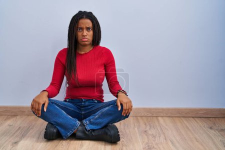 Téléchargez les photos : Jeune afro-américain avec des tresses assises sur le sol à la maison sceptique et nerveux, fronçant les sourcils bouleversés à cause de problèmes. personne négative. - en image libre de droit