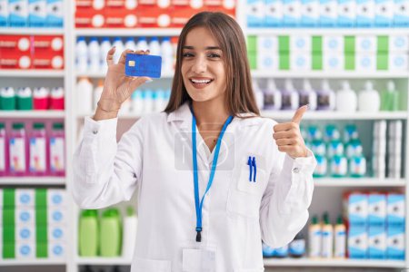 Téléchargez les photos : Jeune femme brune travaillant à la pharmacie pharmacie tenant la carte de crédit souriant heureux et positif, pouce levé faire excellent et signe d'approbation - en image libre de droit