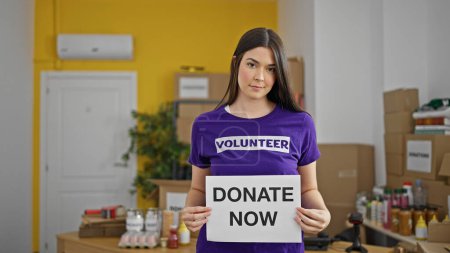 Foto de Joven hermosa hispana voluntaria sosteniendo donar ahora mensaje en el centro de caridad - Imagen libre de derechos