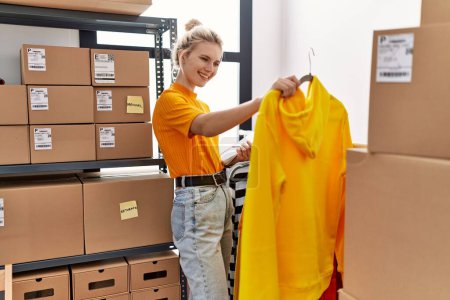 Téléchargez les photos : Jeune femme blonde ecommerce ouvrier d'entreprise organisant des vêtements sur porte-manteau au bureau - en image libre de droit