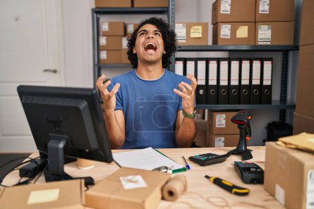 Photo for Hispanic man with curly hair working at small business ecommerce crazy and mad shouting and yelling with aggressive expression and arms raised. frustration concept. - Royalty Free Image