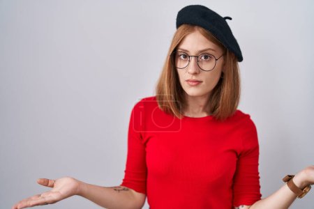 Téléchargez les photos : Jeune femme rousse debout portant des lunettes et un béret désemparé et confus avec les bras ouverts, aucune idée concept. - en image libre de droit