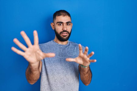 Téléchargez les photos : Middle east man with beard standing over blue background afraid and terrified with fear expression stop gesture with hands, shouting in shock. panic concept. - en image libre de droit
