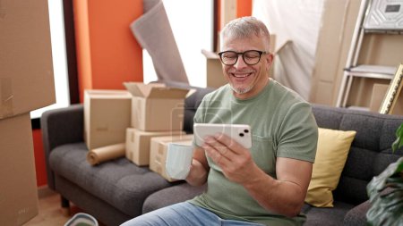 Foto de Hombre de pelo gris de mediana edad viendo vídeo en el teléfono inteligente bebiendo café en un nuevo hogar - Imagen libre de derechos