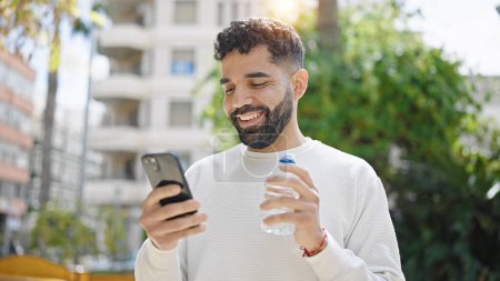 Téléchargez les photos : Jeune homme hispanique tenant une bouteille d'eau en utilisant un smartphone au parc - en image libre de droit