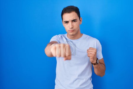 Téléchargez les photos : Jeune homme hispanique debout sur fond bleu coups de poing poing pour se battre, attaque agressive et en colère, menace et violence - en image libre de droit