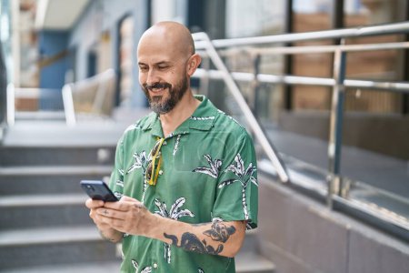 Téléchargez les photos : Jeune homme chauve souriant confiant en utilisant un smartphone dans la rue - en image libre de droit
