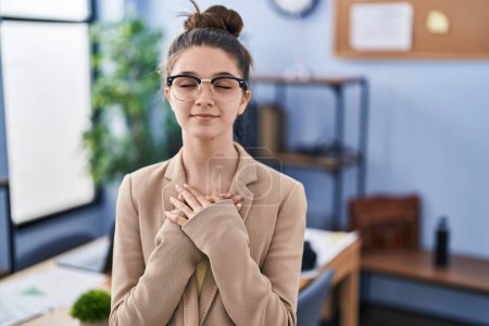 Photo for Teenager girl working at the office wearing glasses smiling with hands on chest with closed eyes and grateful gesture on face. health concept. - Royalty Free Image
