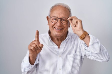 Foto de Senior man with grey hair holding glasses with hand surprised with an idea or question pointing finger with happy face, number one - Imagen libre de derechos