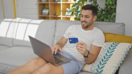 Foto de Joven hombre hispano de compras con portátil y tarjeta de crédito sentado en el sofá en casa - Imagen libre de derechos