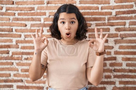 Photo for Young hispanic woman standing over bricks wall looking surprised and shocked doing ok approval symbol with fingers. crazy expression - Royalty Free Image