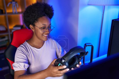 Foto de Mujer afroamericana streamer sonriendo confiado sosteniendo auriculares en la sala de juegos - Imagen libre de derechos