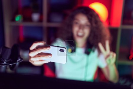 Foto de Young beautiful hispanic woman streamer smiling confident make selfie by smartphone at gaming room - Imagen libre de derechos