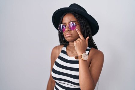 Téléchargez les photos : Jeune afro-américain avec des tresses portant un chapeau et des lunettes de soleil pointant vers l'œil vous regardant geste, expression suspecte - en image libre de droit