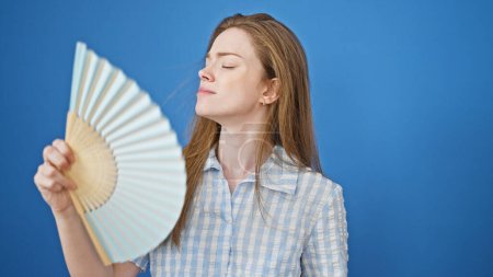 Foto de Mujer rubia joven usando ventilador de mano que sufre por caliente sobre fondo azul aislado - Imagen libre de derechos