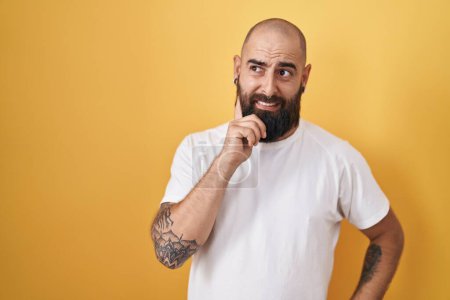 Téléchargez les photos : Jeune homme hispanique avec barbe et tatouages debout sur fond jaune pensant inquiet d'une question, préoccupé et nerveux avec la main sur le menton - en image libre de droit