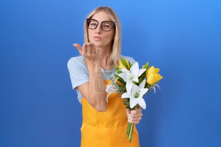 Téléchargez les photos : Jeune femme caucasienne portant tablier de fleuriste tenant des fleurs regardant la caméra souffler un baiser avec la main sur l'air étant belle et sexy. expression d'amour. - en image libre de droit