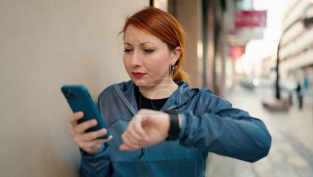 Téléchargez les photos : Jeune rousse femme portant des vêtements de sport en utilisant smartphone regarder chronomètre à la rue - en image libre de droit