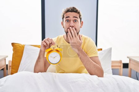 Téléchargez les photos : Hispanic man with beard holding alarm clock in the bed covering mouth with hand, shocked and afraid for mistake. surprised expression - en image libre de droit