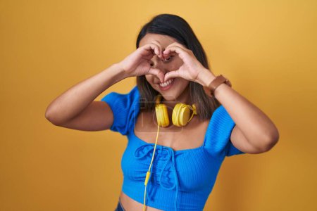 Photo for Hispanic young woman standing over yellow background doing heart shape with hand and fingers smiling looking through sign - Royalty Free Image