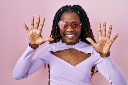 Foto de Mujer africana con el pelo trenzado de pie sobre fondo rosa mostrando y señalando hacia arriba con los dedos número diez mientras sonríe confiado y feliz. - Imagen libre de derechos