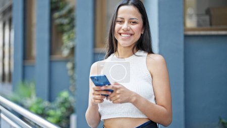 Téléchargez les photos : Jeune belle femme hispanique utilisant smartphone souriant à la rue - en image libre de droit