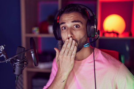 Téléchargez les photos : Hispanic young man playing video games wearing headphones covering mouth with hand, shocked and afraid for mistake. surprised expression - en image libre de droit