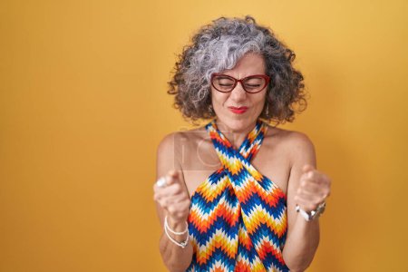 Foto de Middle age woman with grey hair standing over yellow background excited for success with arms raised and eyes closed celebrating victory smiling. winner concept. - Imagen libre de derechos