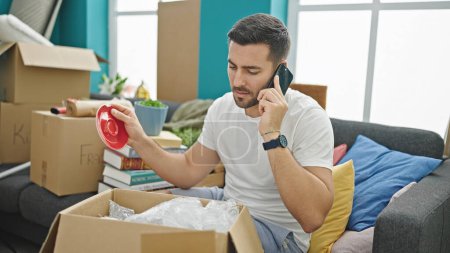 Photo for Young hispanic man unpacking cardboard box talking on smartphone at new home - Royalty Free Image