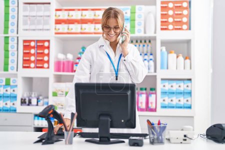 Photo for Young blonde girl pharmacist talking on smartphone using computer at pharmacy - Royalty Free Image